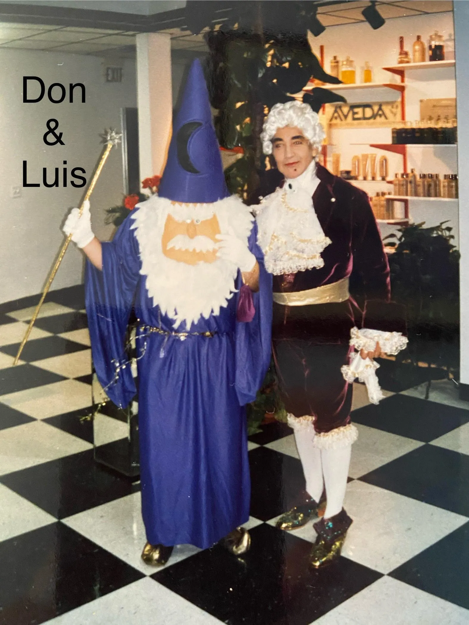 Archival costume party photo of L.A. Monroe co-founders Don, dressed as a wizard in a blue robe, and Luis, in a velvet aristocrat costume, on the checkered salon floor