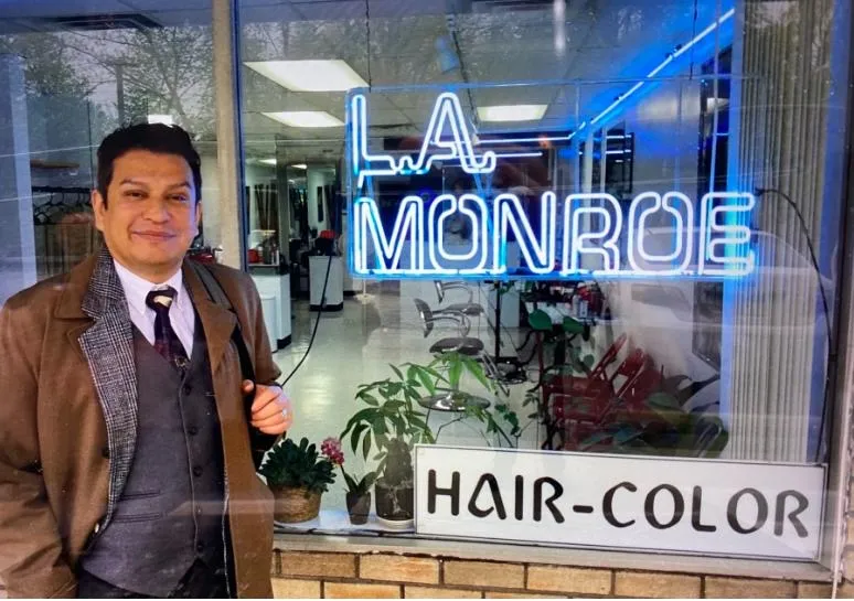 Jesus, stylist at L.A. Monroe Hair Studio, photographed in front of the storefront window with the blue 'L.A. Monroe' neon sign
